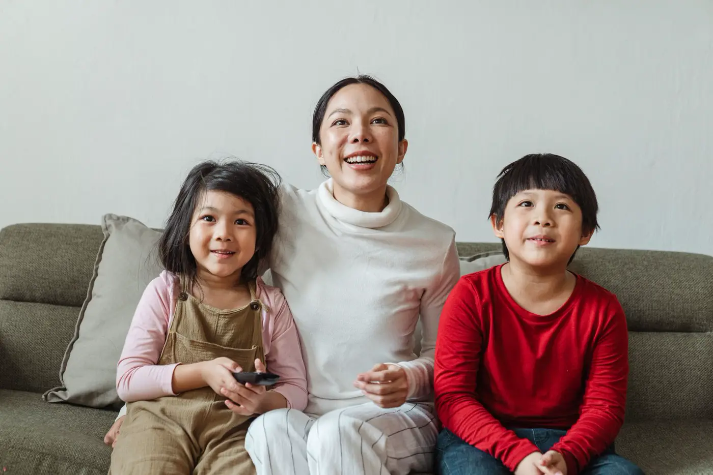A family of four sits on a gray sofa in a living room. One child holds a tablet, and a television is on across the room.